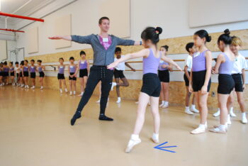 A group of young Introduction to Ballet students in a dance studio wait in line along the barres for their turn. One student copies the teacher's movement, reaching one foot to a tendu side, and both arms outwards, preparing to dance across the floor.