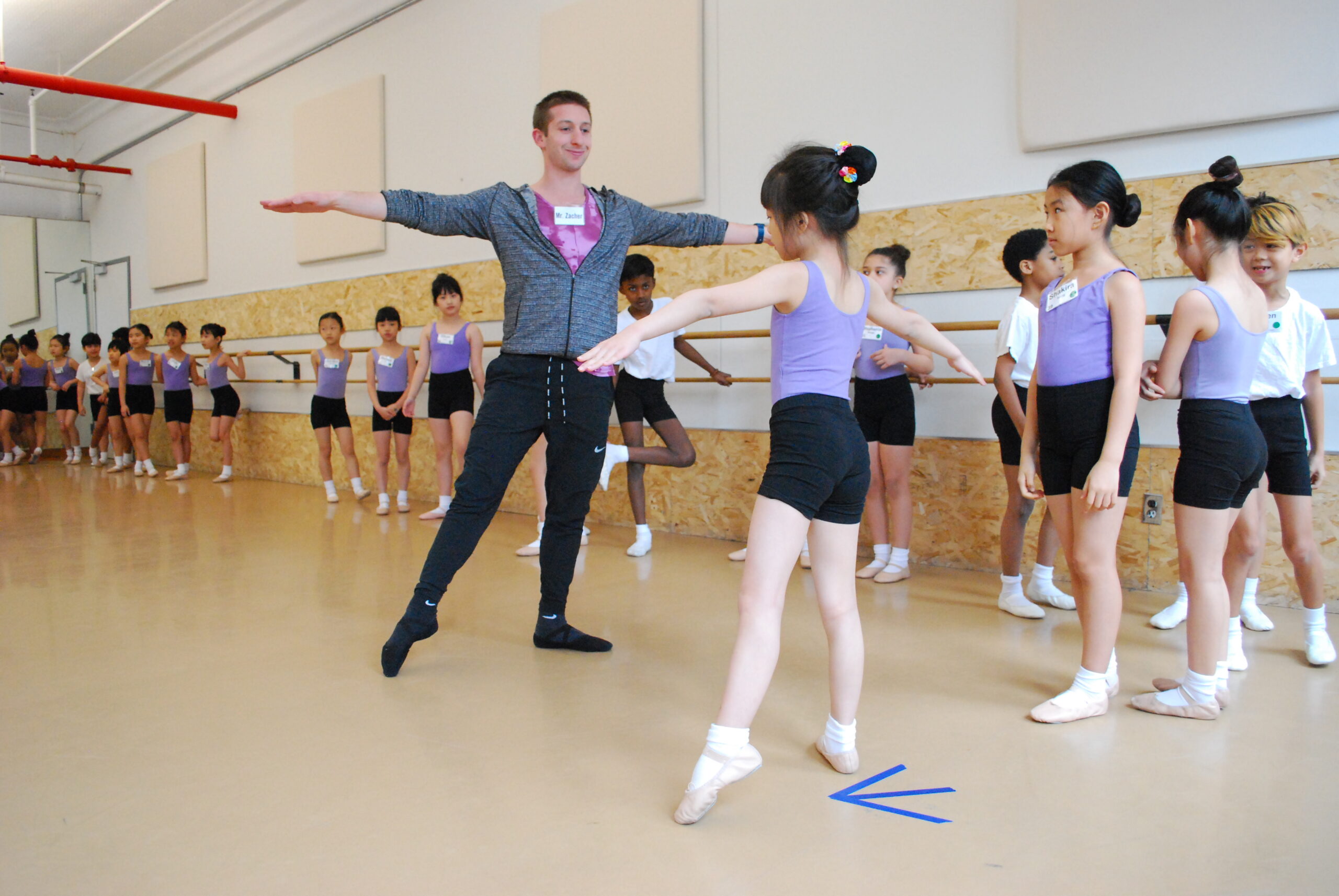 A group of young Introduction to Ballet students in a dance studio wait in line along the barres for their turn. One student copies the teacher's movement, reaching one foot to a tendu side, and both arms outwards, preparing to dance across the floor.