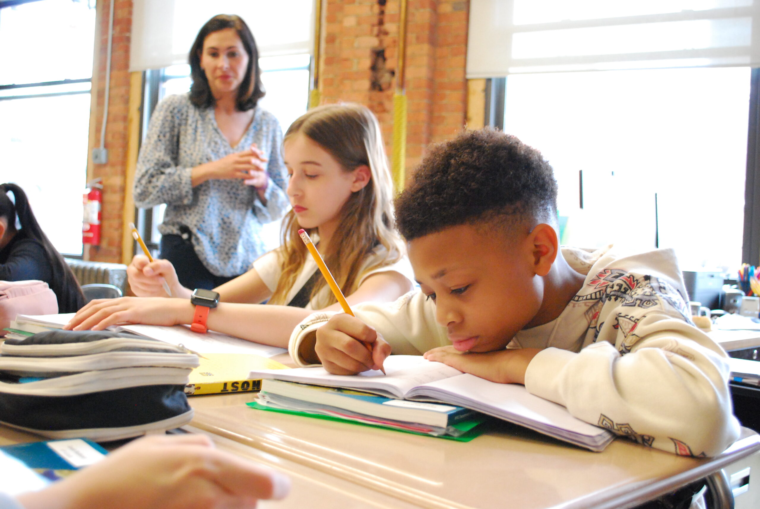 Students in a classroom work at their desks, writing in their notebooks as their teacher walks around and looks on.