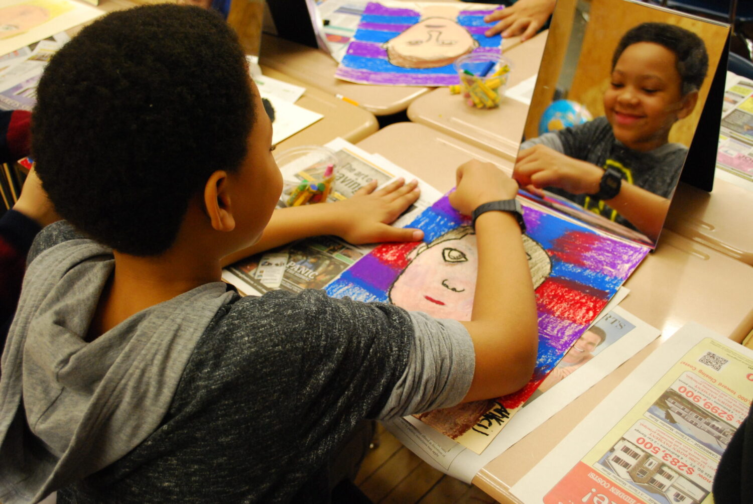 A close up of a visual arts class where students are drawing pastel portraits. Taken from behind, the photo shows a young boy smiling in his reflection from the standing mirror on his desk, while he colors and draws.