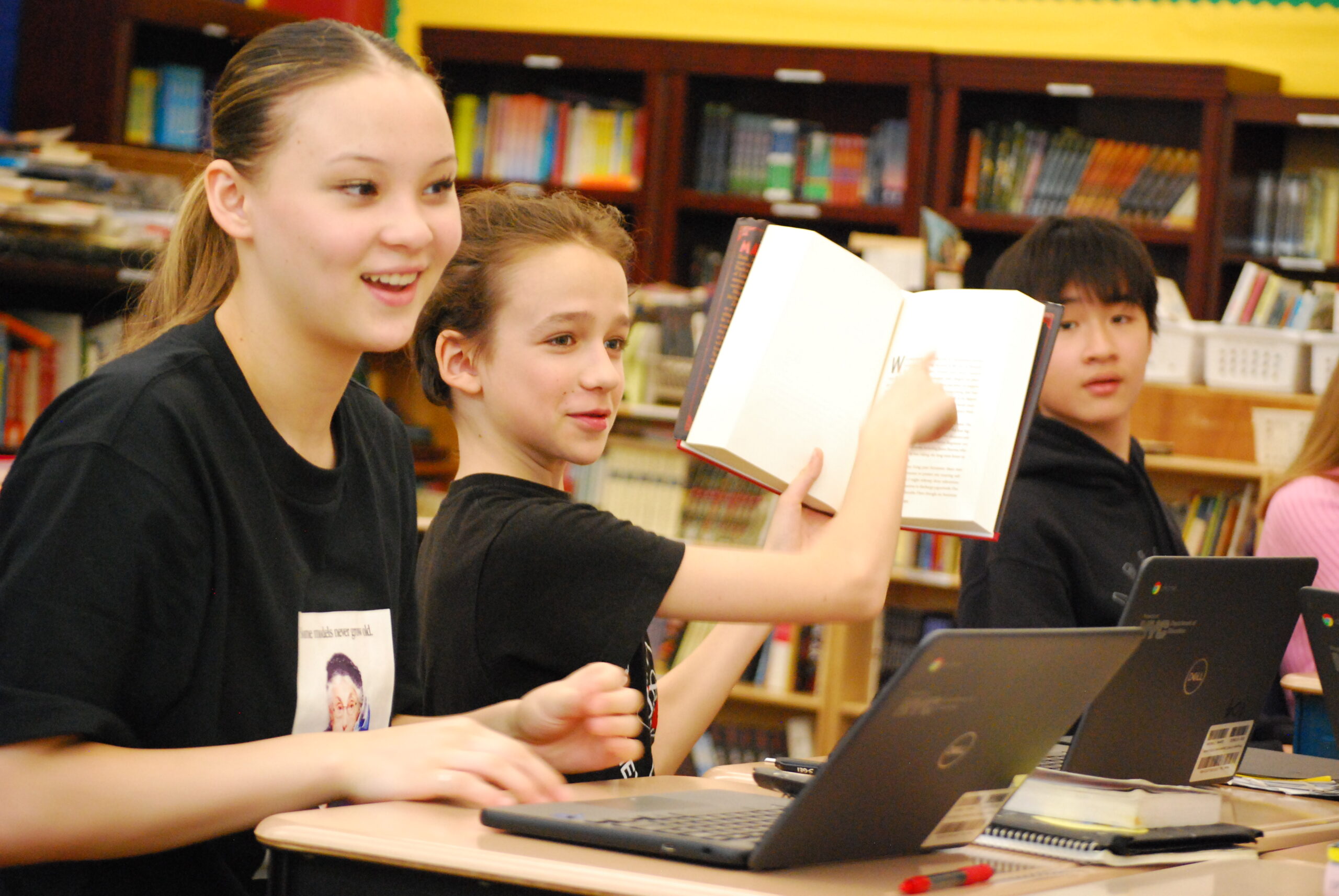 Students in an ELA classroom smile at their desks. One student holds up an open book and points to a page.