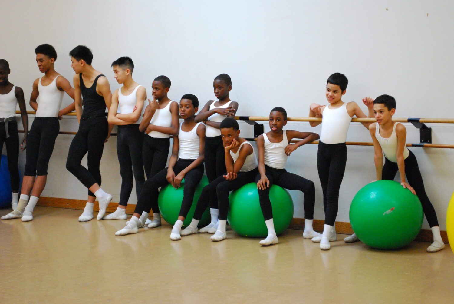 A group of young dancers in white leotards and black tights lean against the barre in the ballet studio in rehearsal. Some of them sit on green Pilates balls.