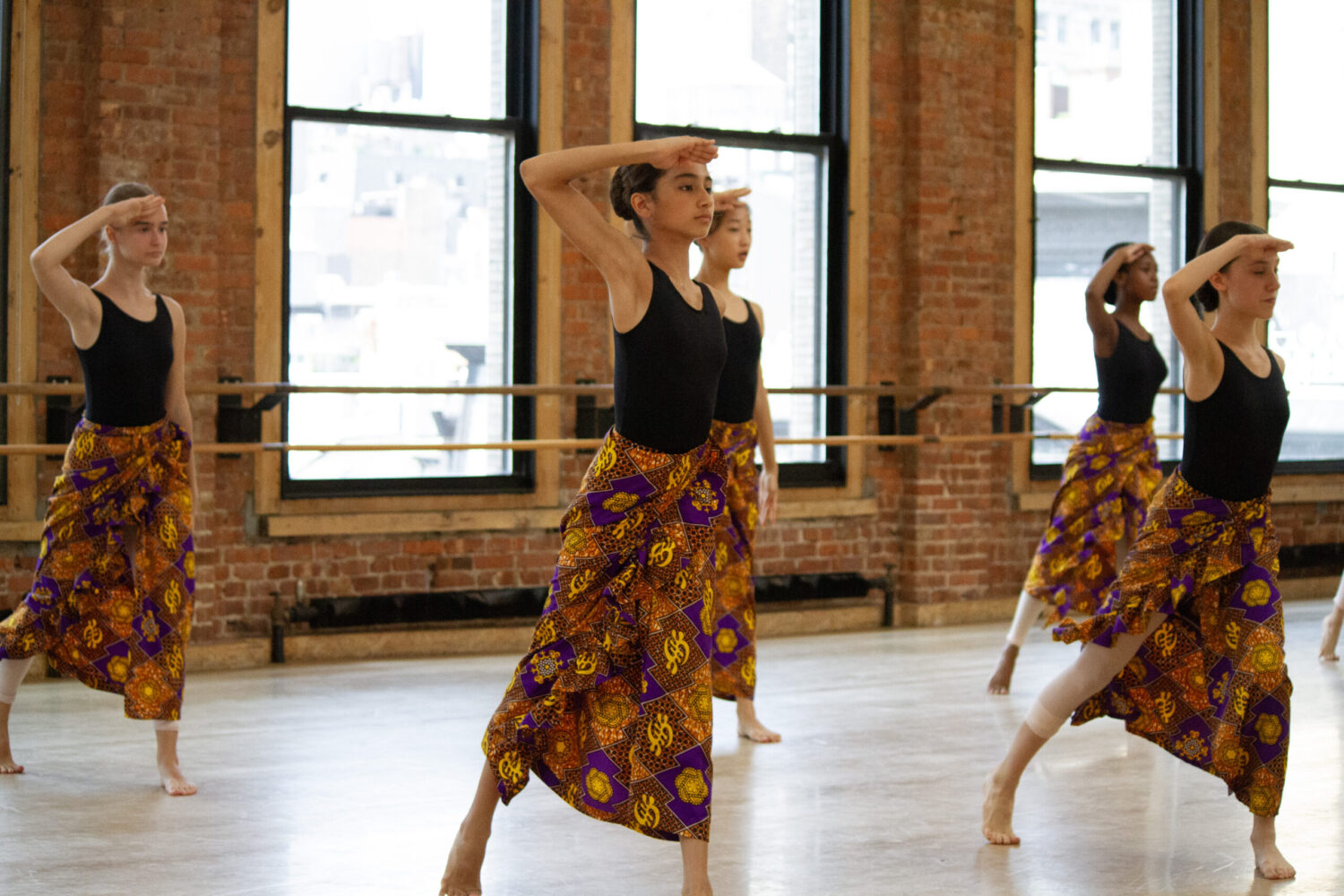 A group of students in a dance studio wear leotards with West African lapa skirts. They lunge forward on one leg, and touch one hand to their foreheads.