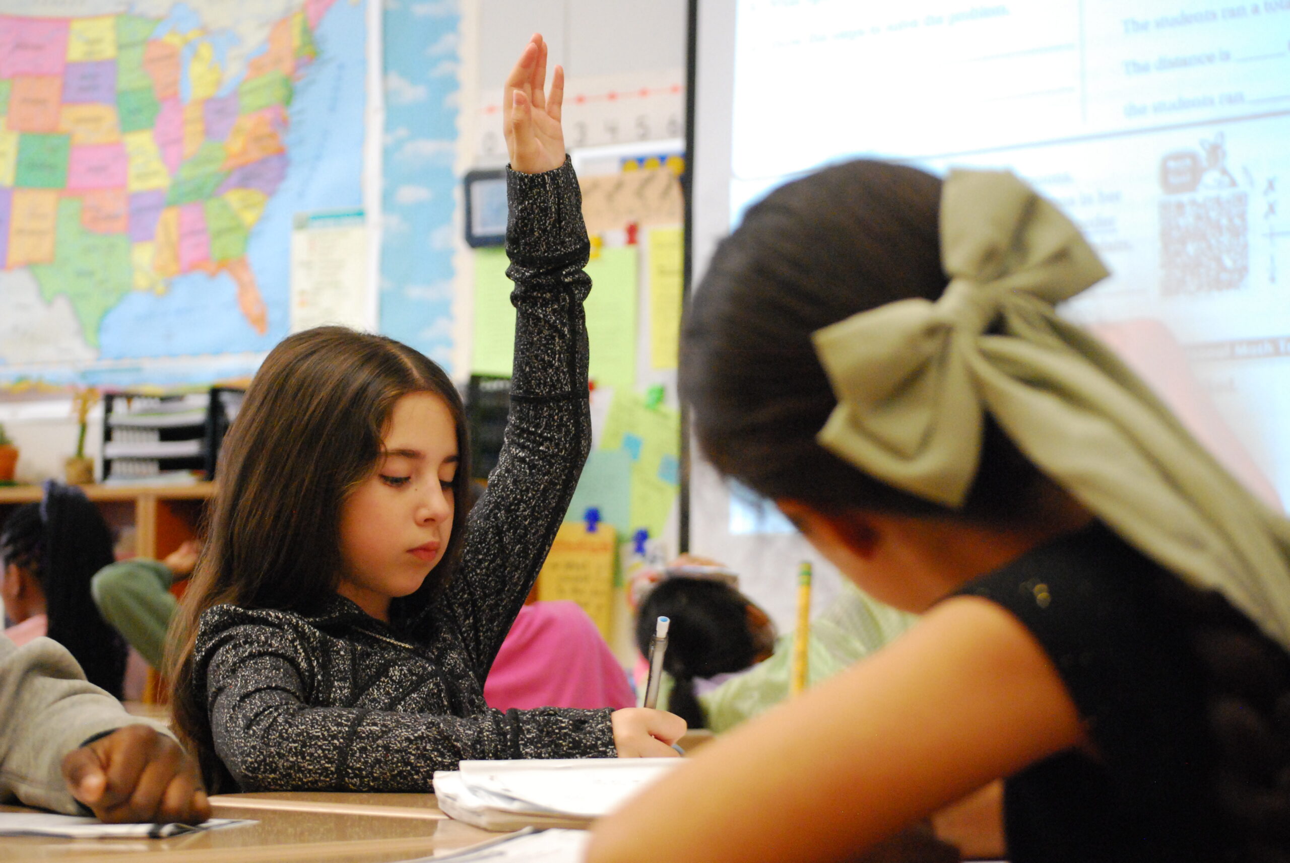 A young girl sits in a classroom working at her desk, raising her hand.