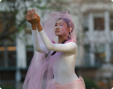 A teenage dancer wearing a white costume with a pink veil-like headpiece gestures forward with both arms, hands cupped together.