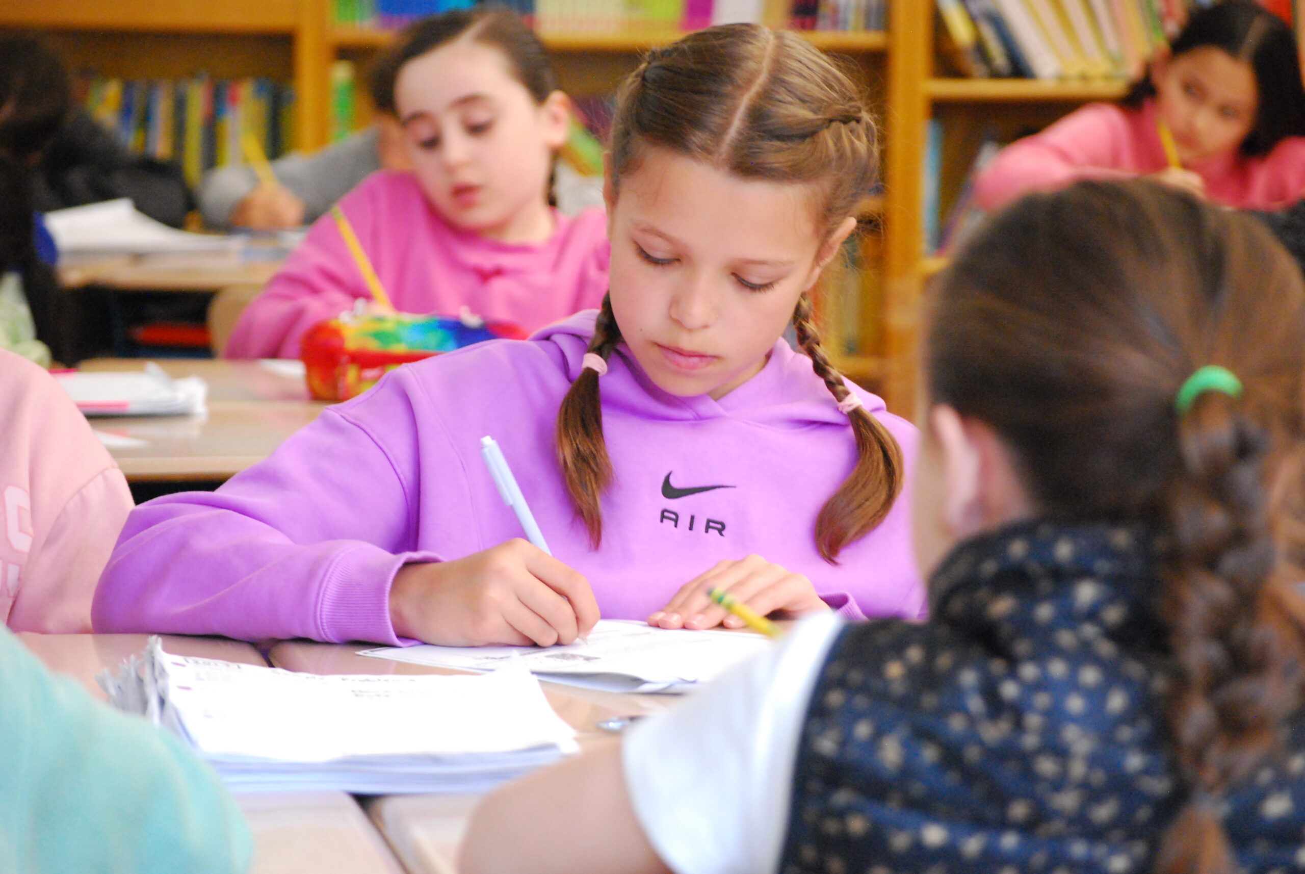 A young student in a classroom works at her desk, writing with a pencil, with other students working in the background.