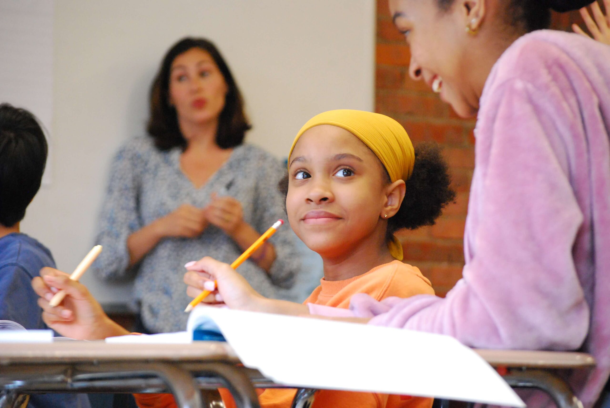 A group of students smile in a classroom, sitting at their desks and holding their pencils while their teacher lectures in the background.