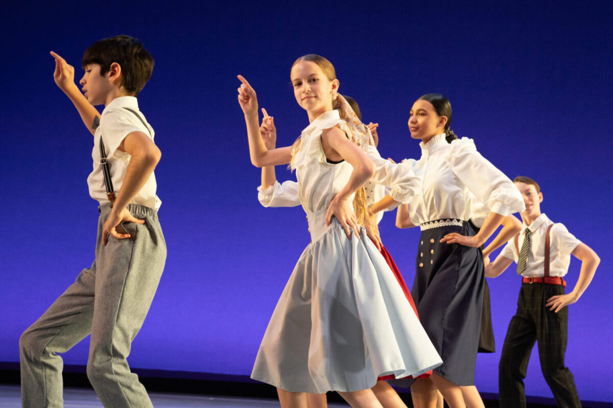 A group of young dancers performs on stage against a blue backdrop. They are dressed in vintage-inspired attire: white blouses and dress pants or skirts. Each dancer strikes a pose with one hand on their hip and the other in the air, showcasing synchronized movements.