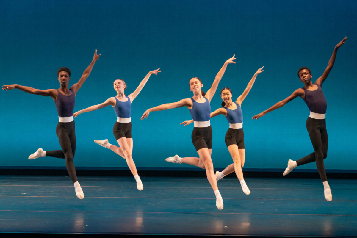Five ballet dancers perform synchronized jumps on stage, each wearing a blue leotard and black shorts, against a blue gradient backdrop.