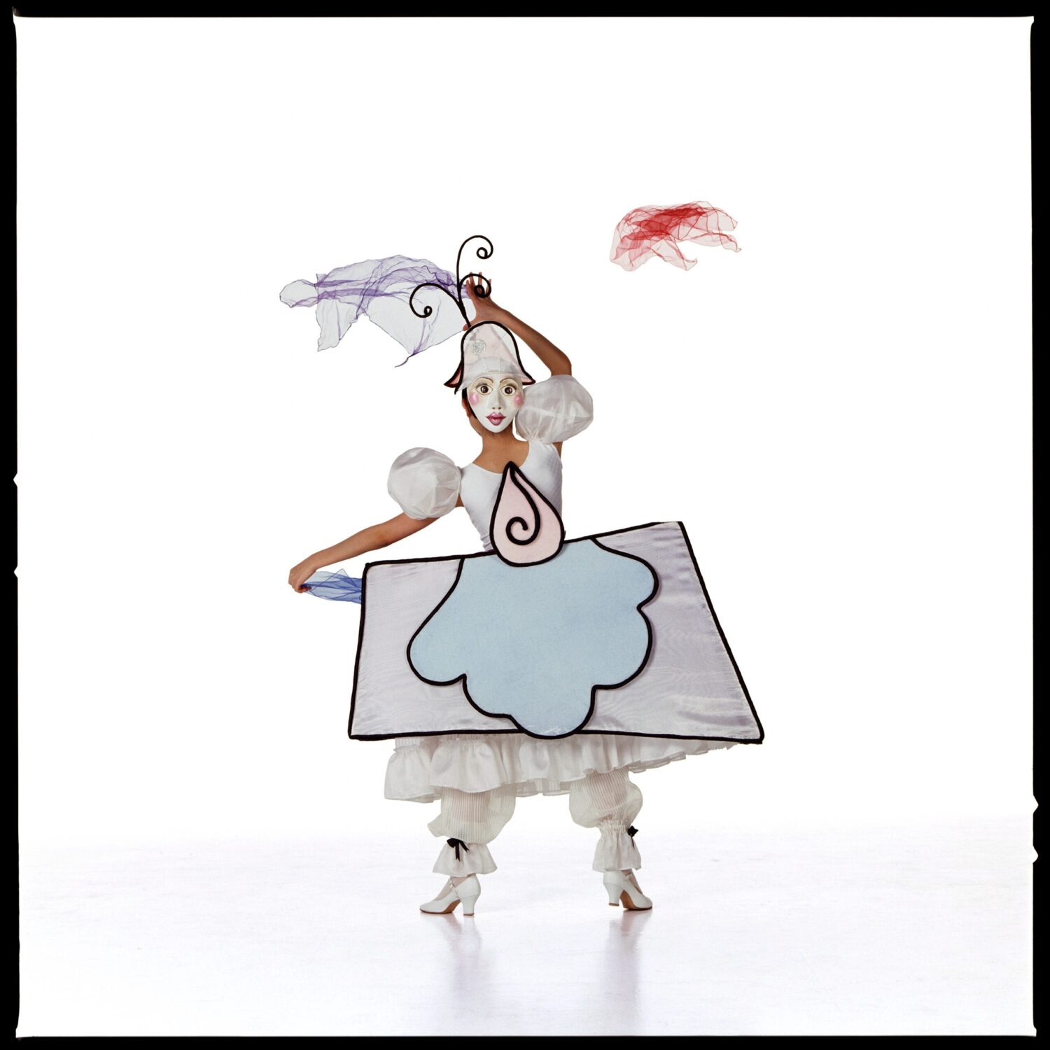A person in a whimsical costume resembling abstract art poses against a white background. The costume includes geometric shapes and delicate, colorful accessories.