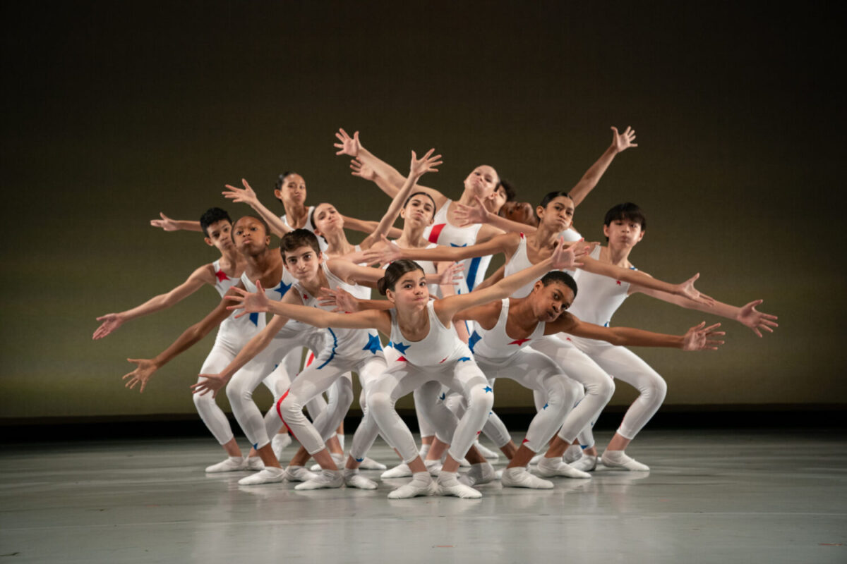 A group of young dancers dressed in white unitards with blue and red stars create a tableau on stage. They form an intricate, visually dynamic pose with outstretched arms and expressive gestures and facial expressions, conveying a sense of unity and movement against a plain backdrop.