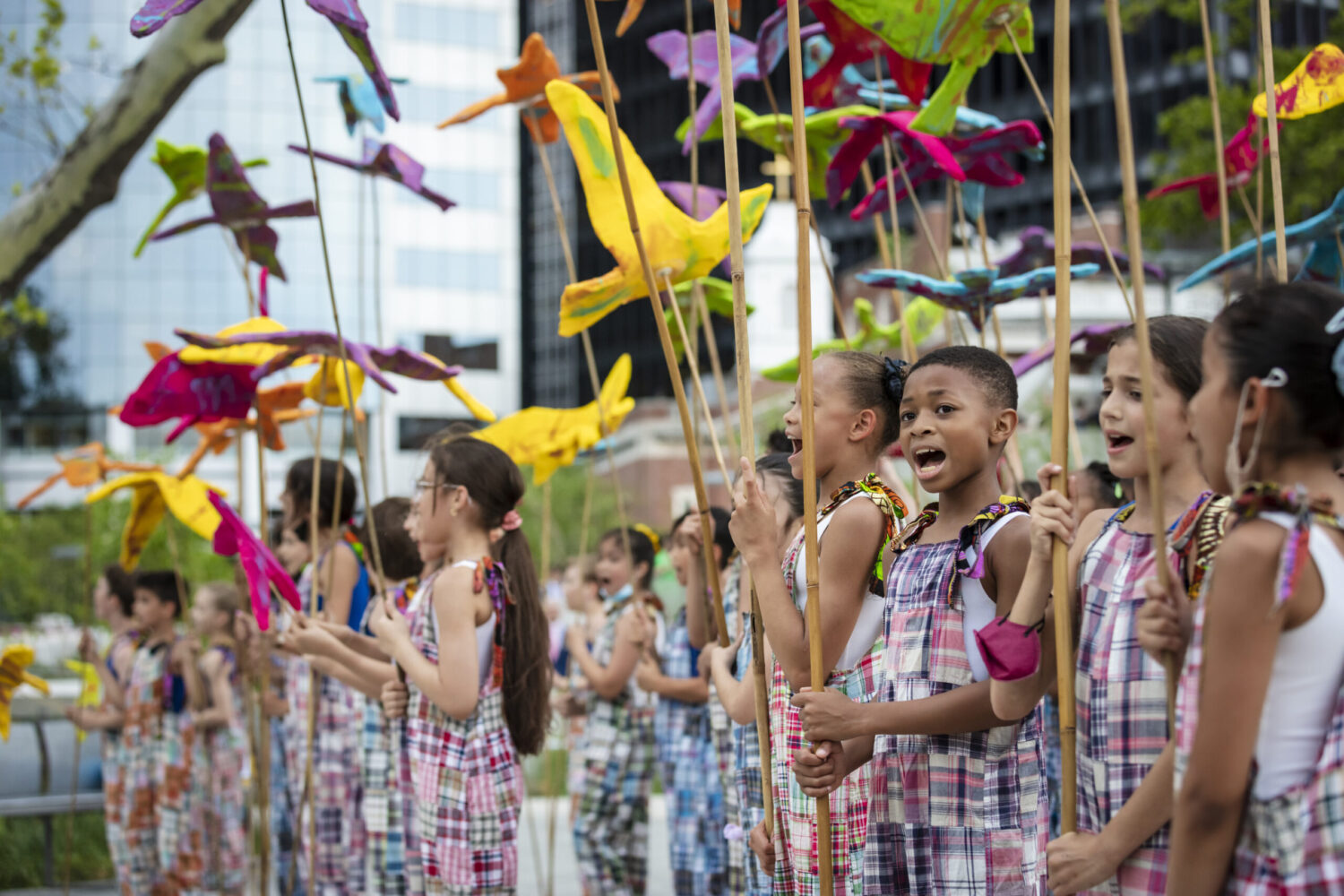 A group of children in colorful, checkered outfits are standing in a row, holding vibrant, bird-shaped kites on long sticks. They are outdoors in an urban setting with trees and buildings in the background, smiling and singing.