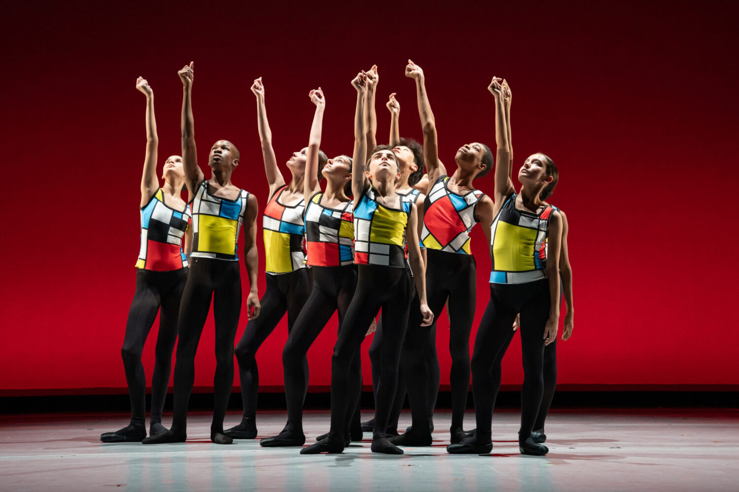 A group of young dancers stands in a formation on stage, wearing colorful geometric-patterned tops inspired by Mondrian's art. They all point upwards with one arm, snapping their finger against a vibrant red background, creating a striking visual contrast.