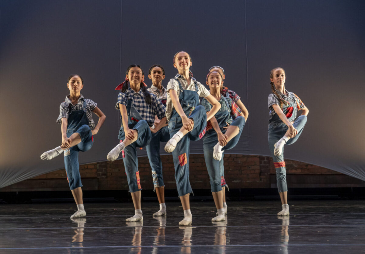 A group of six young dancers, dressed in matching blue overalls and checkered shirts, perform a synchronized routine on stage. They are mid-movement with one leg raised in the air, smiling joyfully in a well-lit, minimalistic set with a brick wall background.