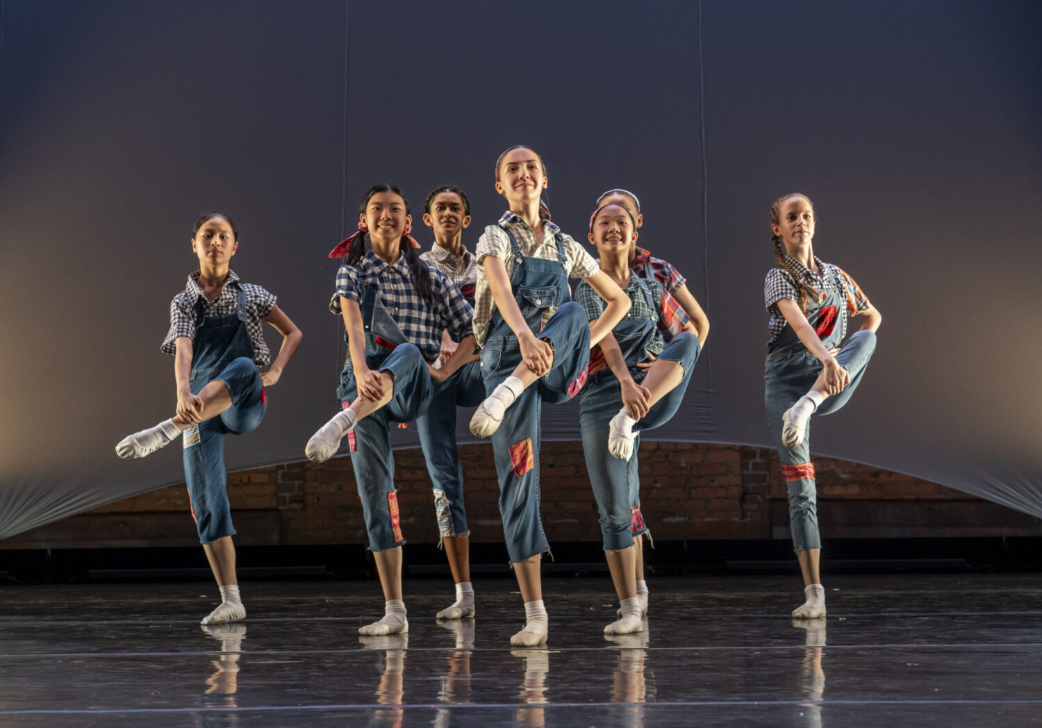 A group of six young dancers, dressed in matching blue overalls and checkered shirts, perform a synchronized routine on stage. They are mid-movement with one leg raised in the air, smiling joyfully in a well-lit, minimalistic set with a brick wall background.