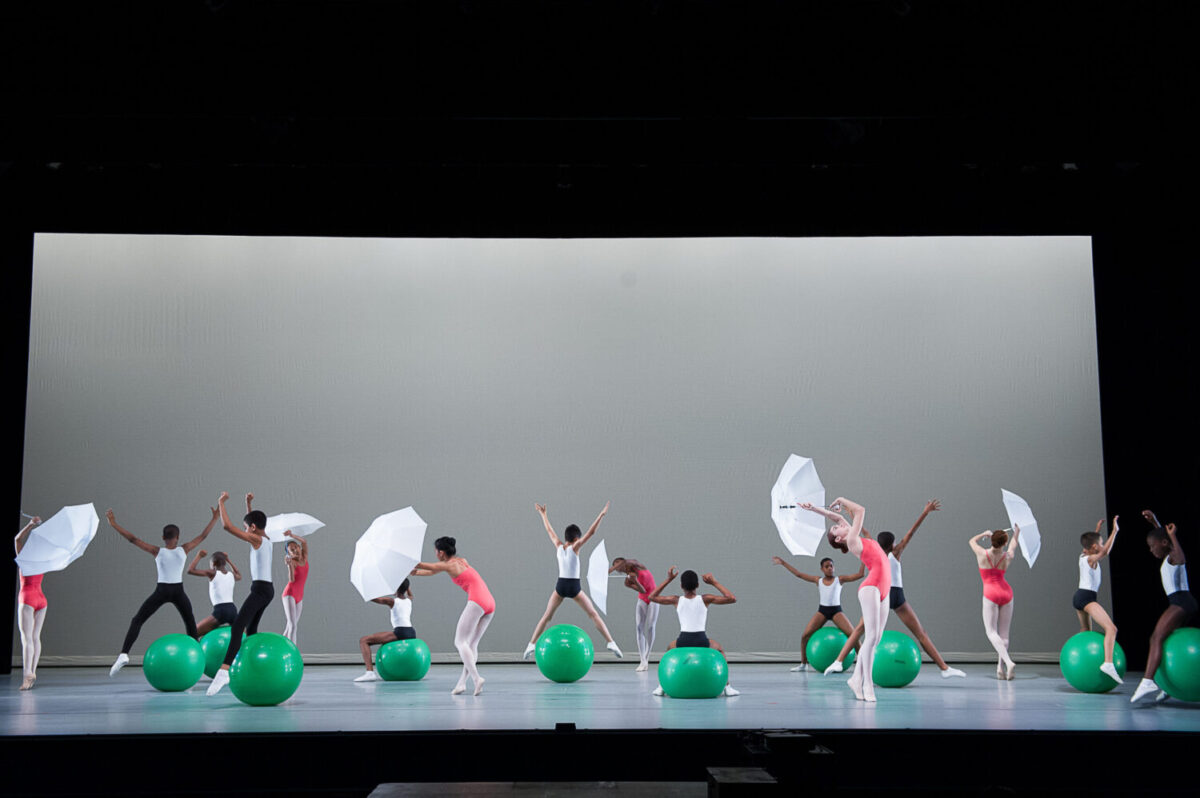 Dancers perform on stage with large green exercise balls and white umbrellas. They are arranged in a broad formation, some standing and others seated on the balls, all mid-movement. The background is a minimalist white while the stage is illuminated brightly.