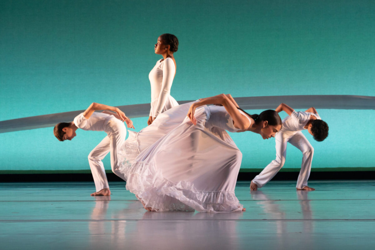 Four dancers dressed in white perform on a stage with a turquoise backdrop. Three dancers are bent in a forward contraction, and the last stands straight in the center holding the skirt of their dress delicately. The performance exudes grace and harmony.