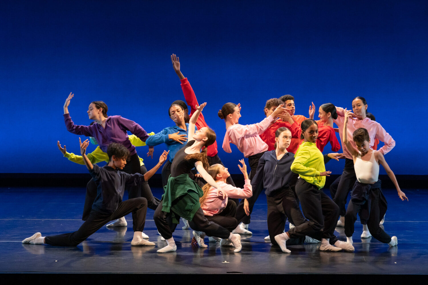 A group of dancers perform on stage against a blue background. They wear colorful sweatsuits with long sleeves in shades of purple, red, yellow, black, green, and blue. The dancers strike dynamic poses with expressive arm movements, creating a visually engaging composition.