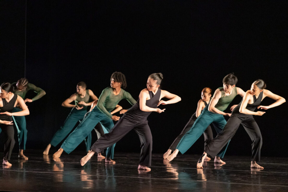 A group of dancers performs on stage, each wearing green and black costumes. They move in unison, holding their hands cupped atop one another with one leg extended to the side against a dark background.