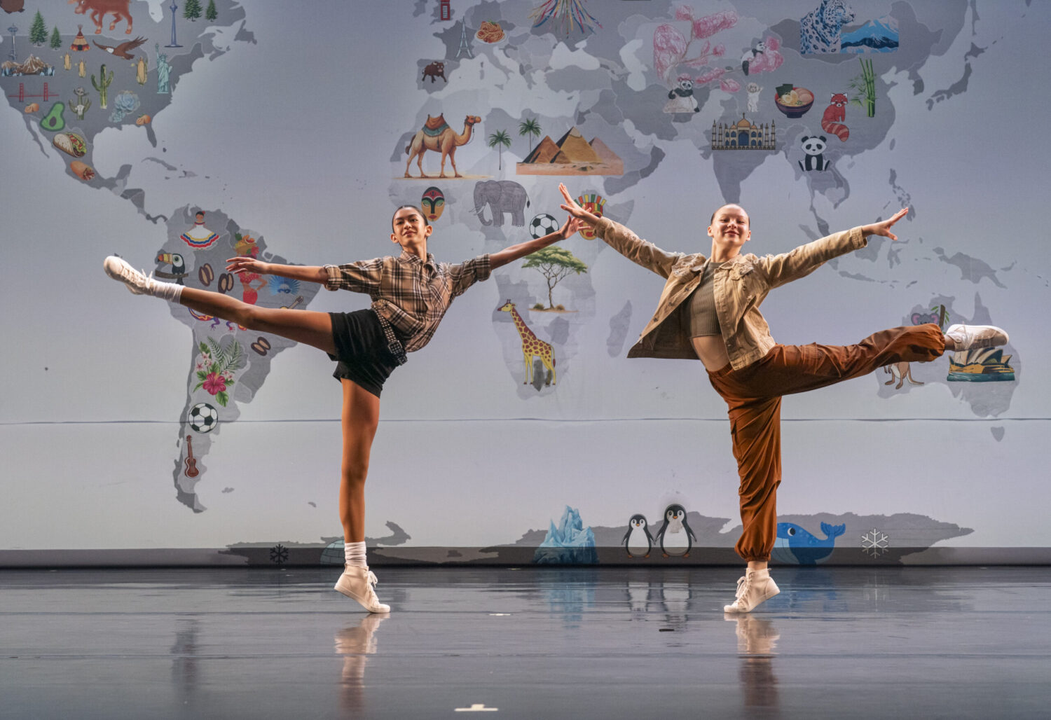 Two ballet dancers are performing on stage with a world map illustrated with animals and landmarks in the background. Striking an arabesque position, the dancer on the left wears a plaid shirt and black shorts, while the dancer on the right wears a beige jacket and orange pants.