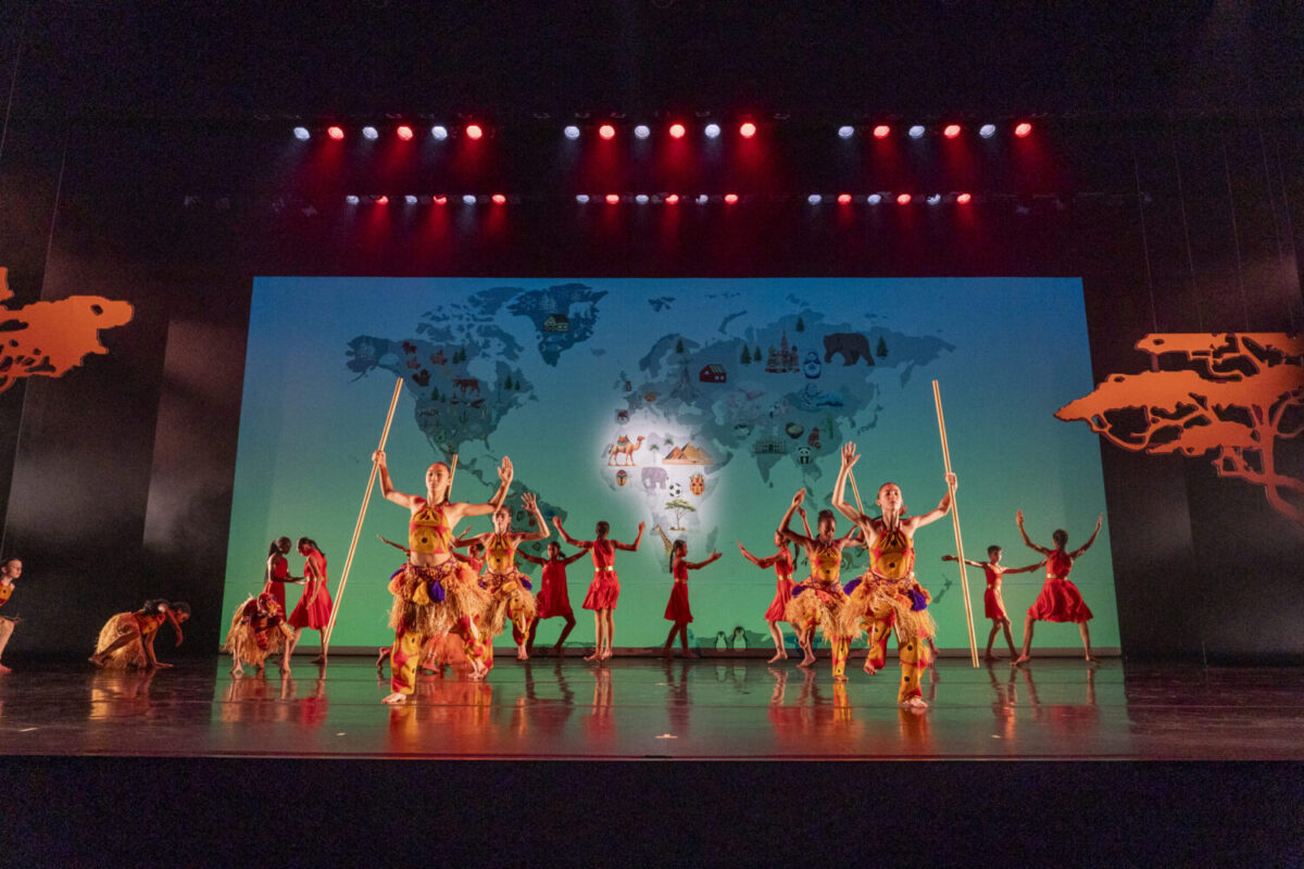 A group of dancers in colorful West African costumes performs on a stage with a backdrop displaying a world map. The stage is lit with red and blue lighting, and dancers hold long staffs while striking dynamic poses. Silhouettes of trees are on each side of the stage.