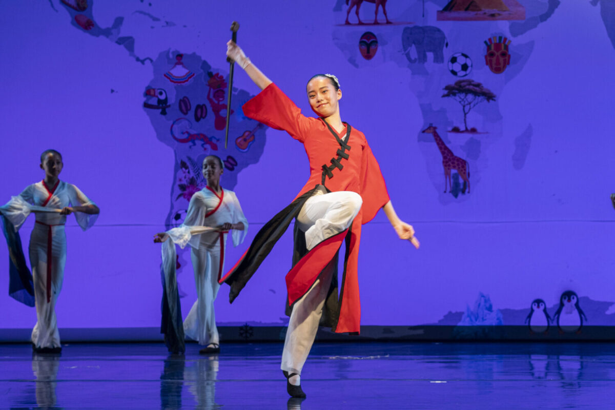 A dancer in traditional Chinese folk dance attire, wearing a red and black dress, performs a dynamic pose on stage with one leg raised while yielding a sword. Three other dancers in white costumes stand in the background. The backdrop features a world map with various cultural icons and images.