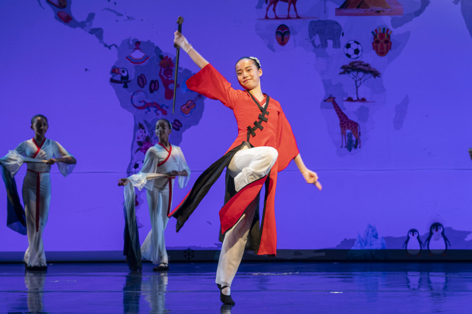 A dancer in traditional Chinese folk dance attire, wearing a red and black dress, performs a dynamic pose on stage with one leg raised while yielding a sword. Three other dancers in white costumes stand in the background. The backdrop features a world map with various cultural icons and images.