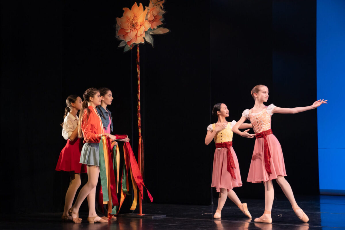 Five young ballet dancers are on stage. Three of them are clustered together to the left, holding colorful ribbons attached to a tall pole topped with a large flower. The other two, dressed in pink costumes, are gesturing towards center stage.