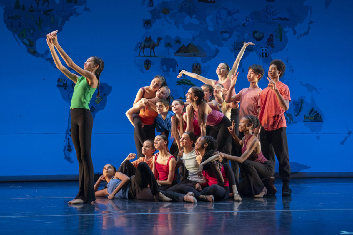 A diverse group of dancers poses joyfully on stage for a selfie. They are dressed in various colorful outfits and are gathered closely together. The background features a world map with animal illustrations, adding a global theme to the lively scene.