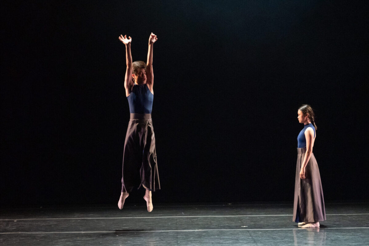 Two dancers performing on stage with a dark background. One dancer, dressed in a blue top and dark pants, is mid-jump with arms extended upward. The other dancer, also in a blue top and dark pants, stands to the right, watching the performance.