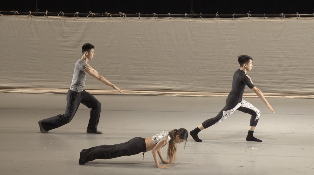 Three dancers dressed in neutral tones are seen on stage performing contemporary movements. The two upstage dancers are in a lunging position with arms extended, while the downstage dancer is in a plank position.