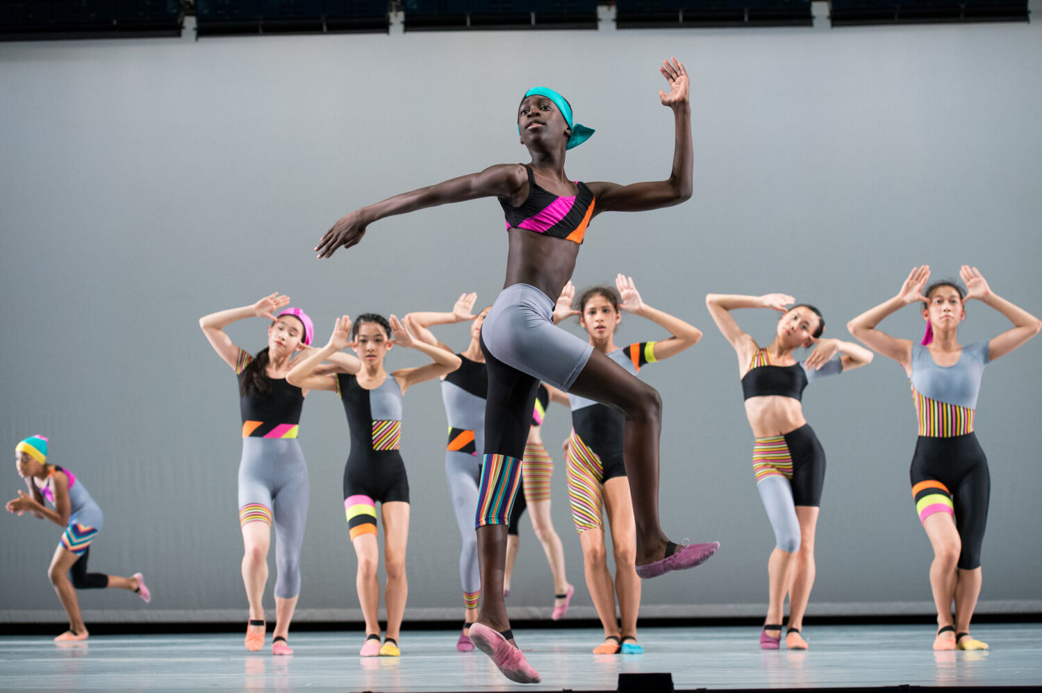 A group of dancers in vibrant, colorful, asymmetrical workout attire perform on stage. The central dancer, leaping gracefully, wears a green headband and coordinated activewear with bright stripes. Other dancers strike synchronized poses in the background against a plain backdrop.