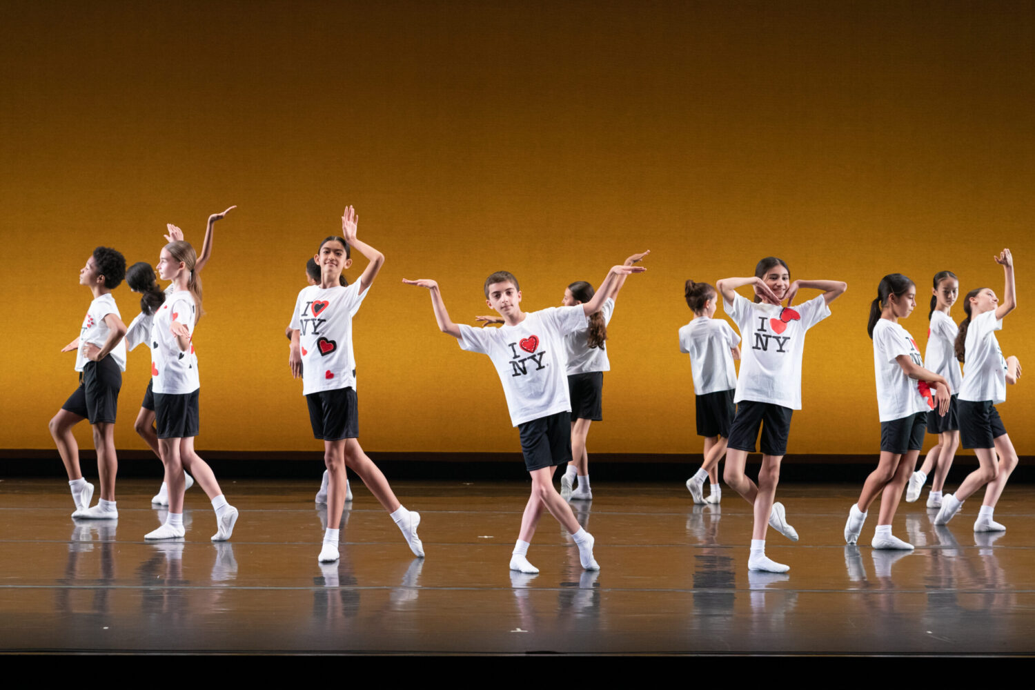 A group of children wearing &quot;I ♥ NY&quot; t-shirts and black shorts perform a dance routine on stage. They strike various expressive poses against an orange backdrop, synchronizing their movements. Some have their arms raised, while others extend arms outward.