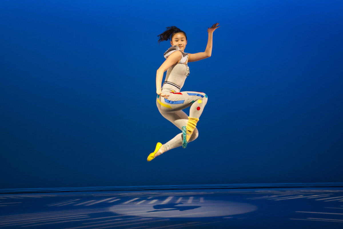 A dancer performs a mid-air leap against a bright blue background. She wears a white outfit with colorful patterns, including a yellow sock, and blue and red accents. Her arm is raised, and she appears joyful and energetic. Shadows cast intricate shapes on the floor.