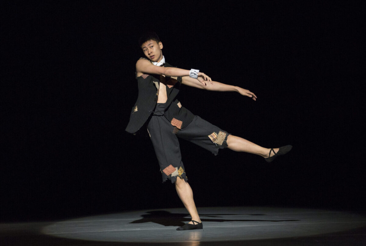 A dancer performs on stage against a dark background, balancing on one leg with the other leg extended to the side. They wear a black, short, sleeveless outfit with patches and a white wristband. The spotlight highlights their precise posture and form.