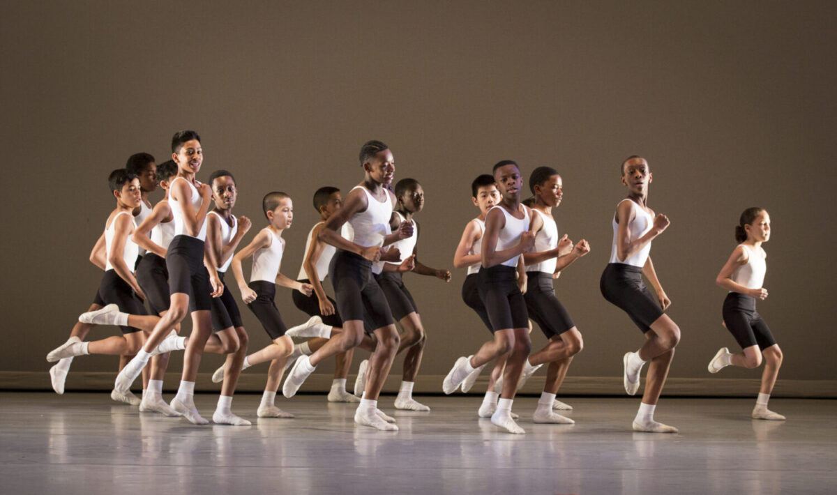 A group of students perform Eliot Feld&#039;s &quot;Quickstep&quot; on stage. Wearing white leotards with black shorts and white ballet slippers, a group of young boys jog in unison.