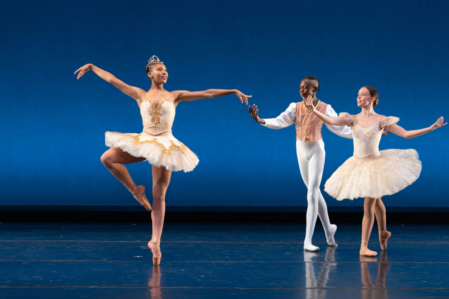 Three ballet dancers perform on stage against a blue backdrop. The soloist, placed center, wears a tiara and a golden tutu while balancing gracefully on one leg. The dancer to the right and the dancer to the left, both in elegant costumes, gesture towards the soloist.
