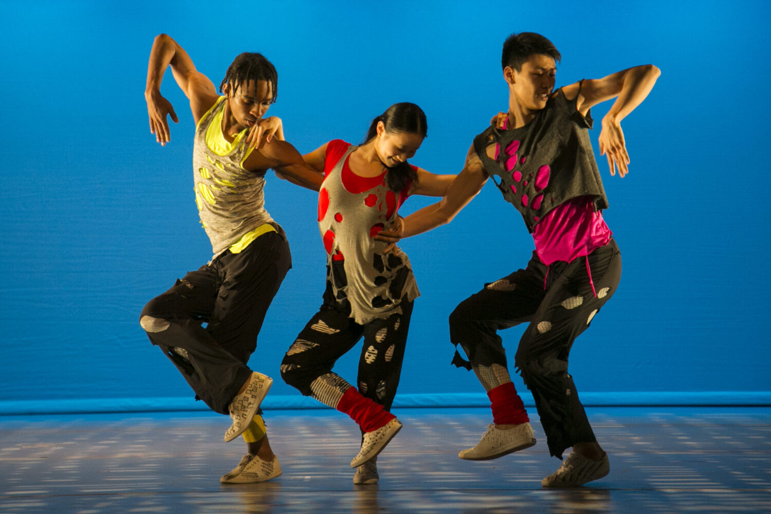 Three dancers perform side by side on stage in vibrant, colorful outfits with dynamic poses. The background is a solid blue, emphasizing their expressive movements. They all wear loose-fitting pants and tops with abstract patterns, blending coordination with individual flair.