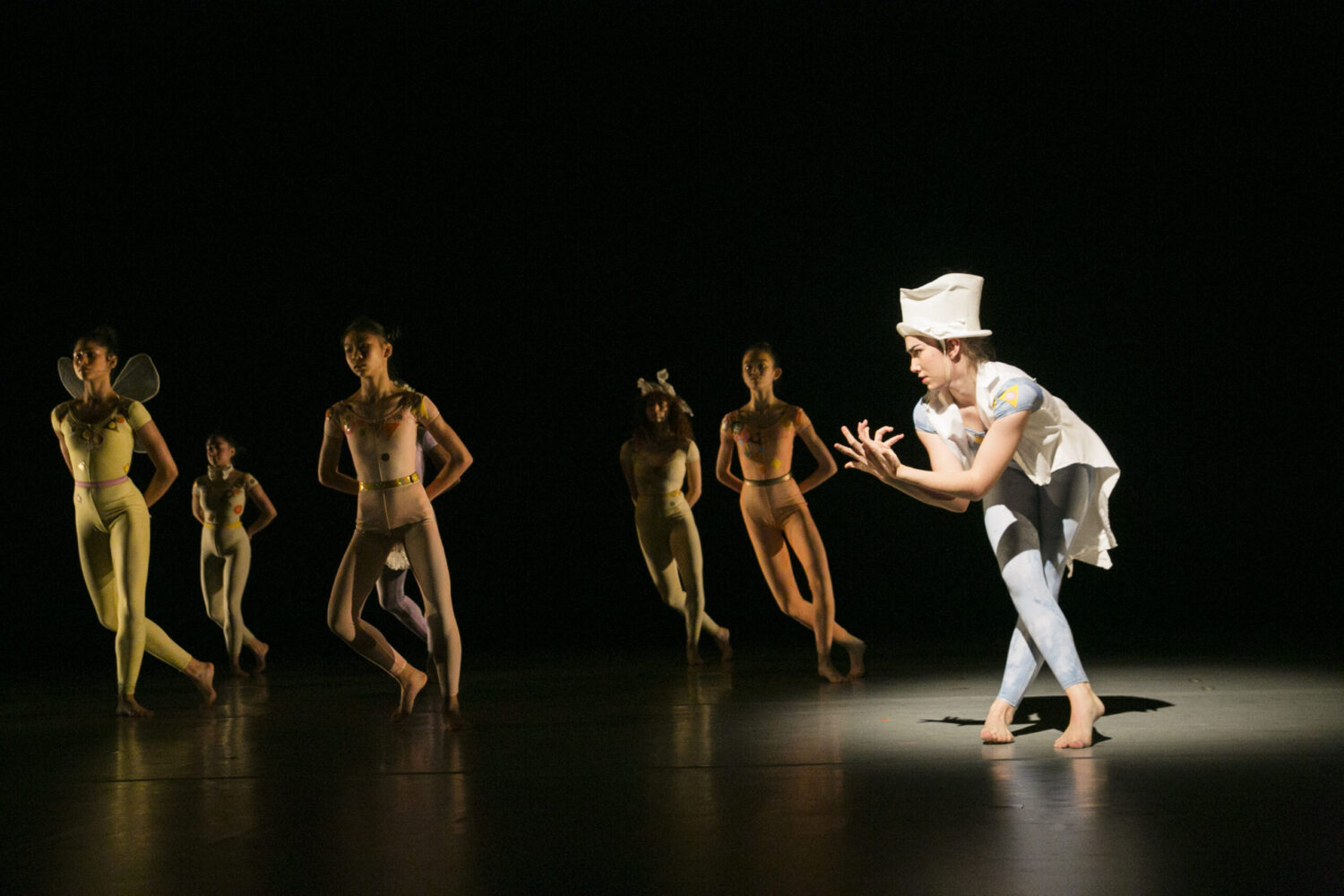 A group of dancers perform on a dimly lit stage. Five dancers in beige costumes move in the background, while a dancer in a white costume and a tall hat is illuminated in the foreground, striking a dramatic pose with outstretched hands.