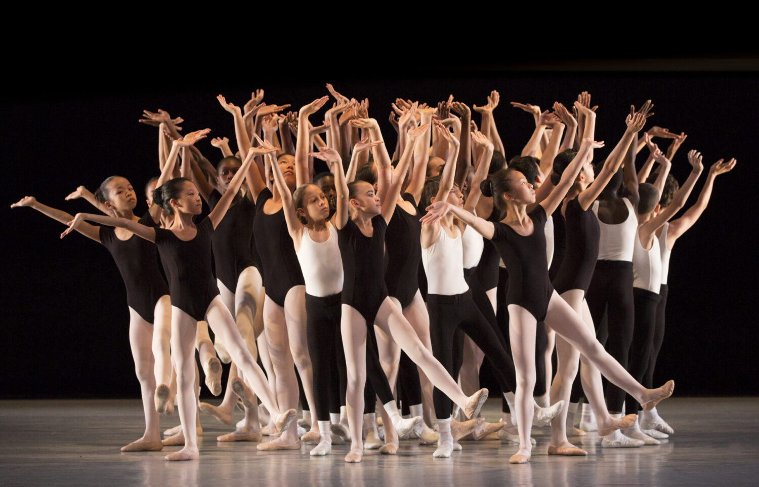 A group of ballet dancers, wearing black leotards and white tights, perform on stage. They are standing close together, arms raised with flexed wrists and feet, creating a dynamic and expressive formation. The background is dark, focusing attention on the dancers.