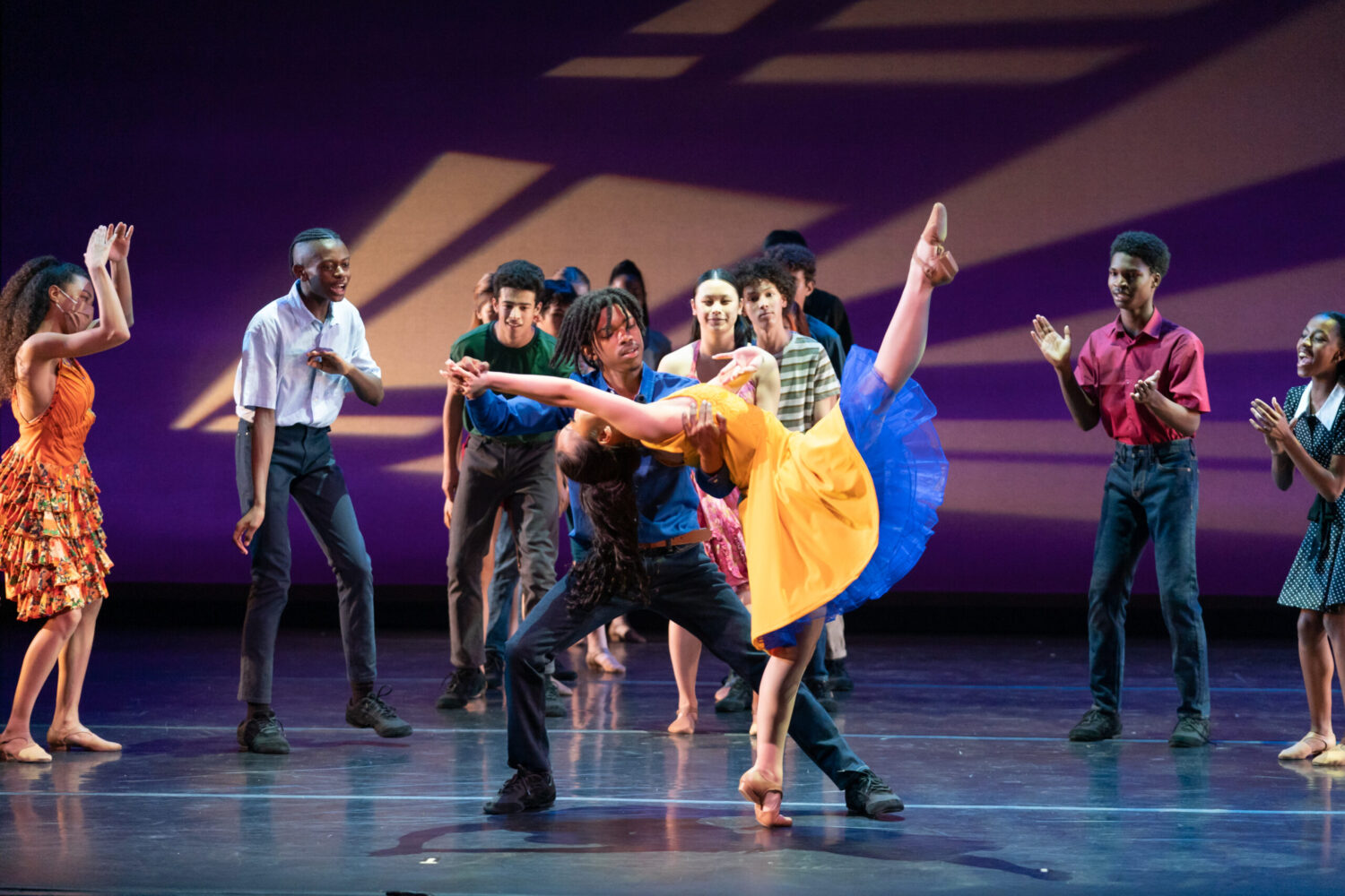 A group of dancers performs on stage with colorful lighting. The ensemble in the background claps and watches as a central pair, consisting of a male and female dancer, execute a dramatic lift and leg extension. The woman wears a yellow dress, and the man supports her.