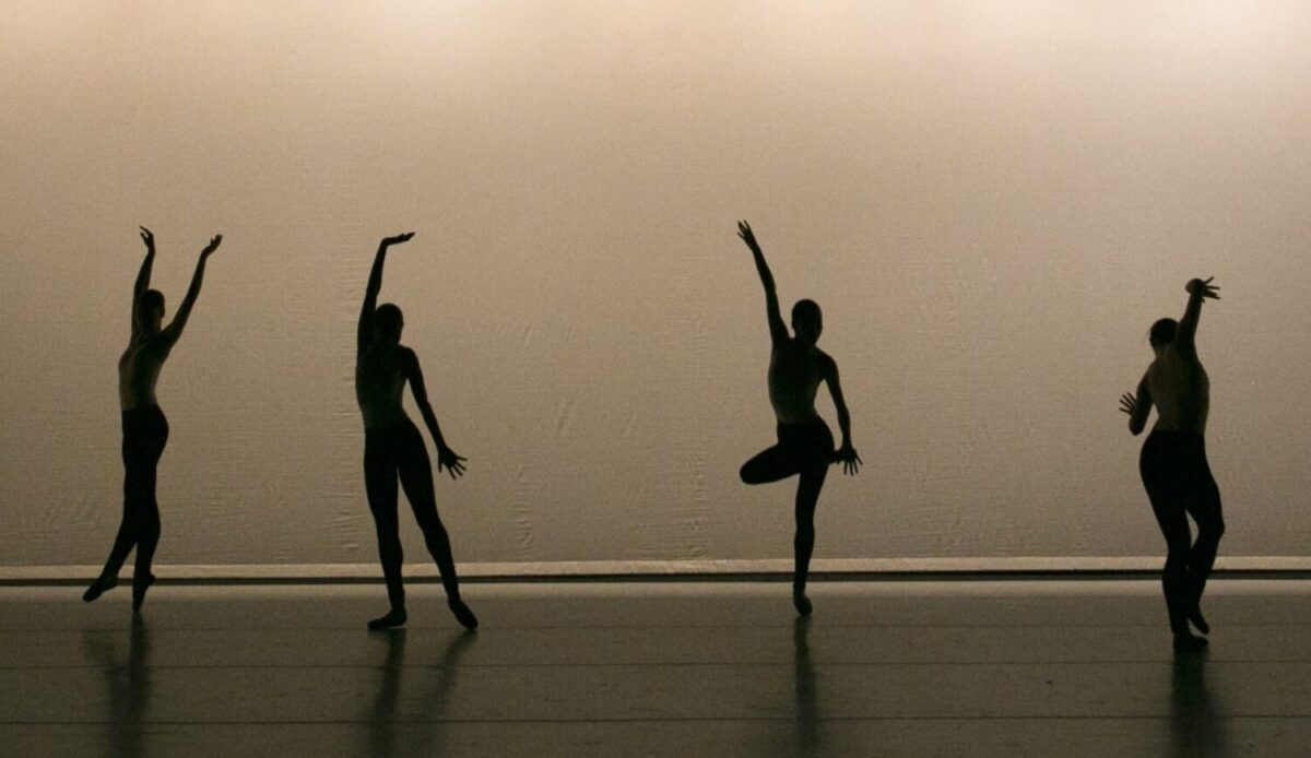 Four dancers wearing pointe shoes are in silhouette on a stage. They dance in a line, moving through graceful balletic gestures.