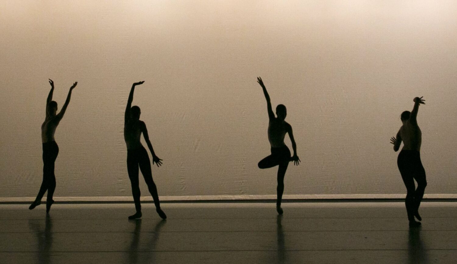 Four dancers wearing pointe shoes are in silhouette on a stage. They dance in a line, moving through graceful balletic gestures.