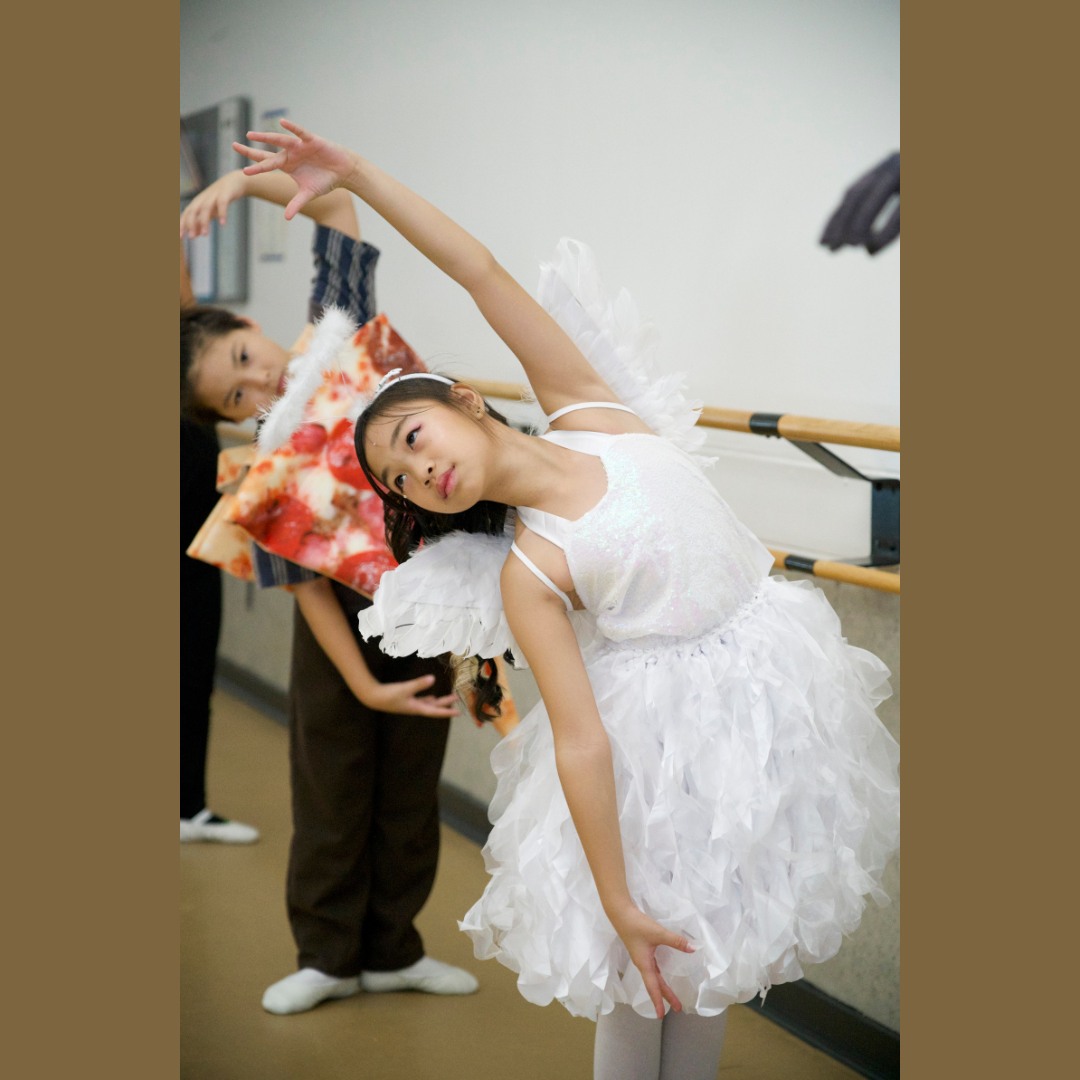 Two young students dressed in an angel and a pizza Halloween costume dance in a ballet class, stretching gracefully away from the barre.