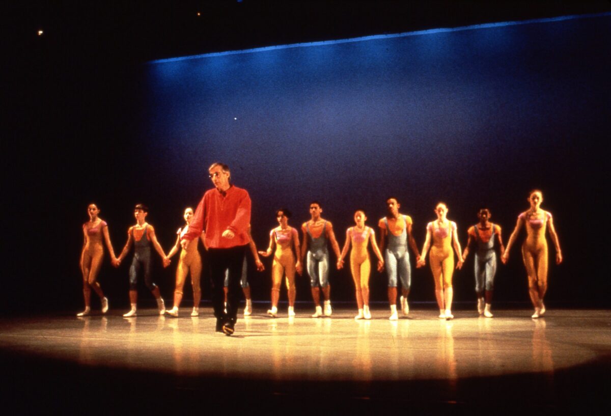 Eliot Feld shows a group of dancers how to bow. The dancers walk in a line holding hands, standing in costume behind him on stage at The Joyce Theater.