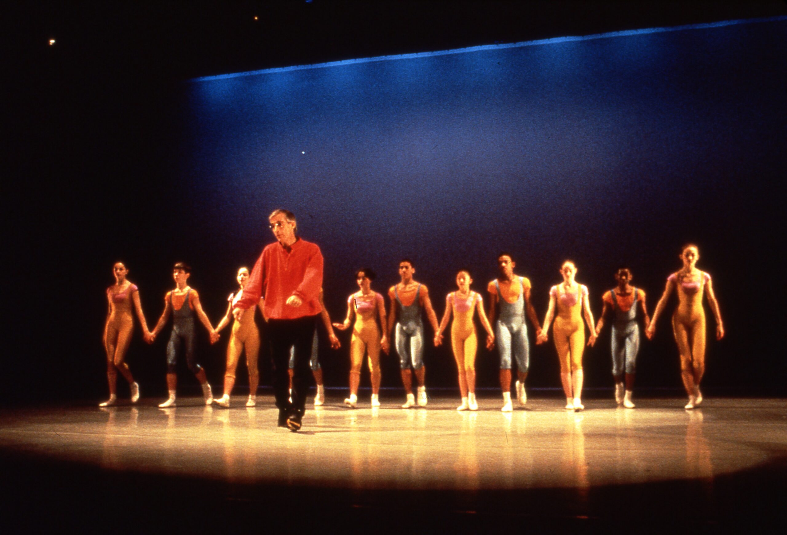 Eliot Feld shows a group of dancers how to bow. The dancers walk in a line holding hands, standing in costume behind him on stage at The Joyce Theater.