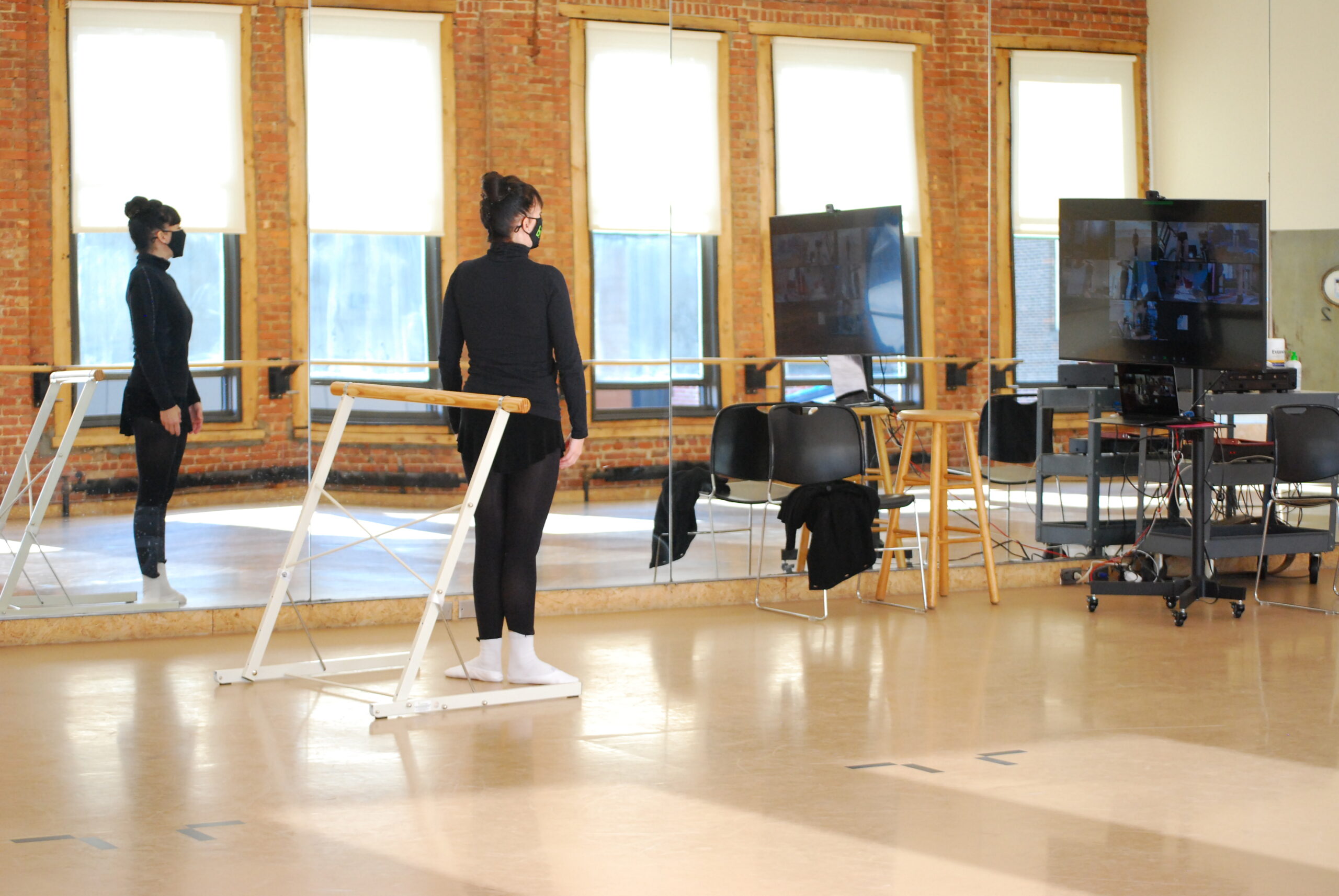 Ashley Tuttle stands in a dance studio in first position at a portable barre, dancing in front of a screen of students taking class on Zoom.