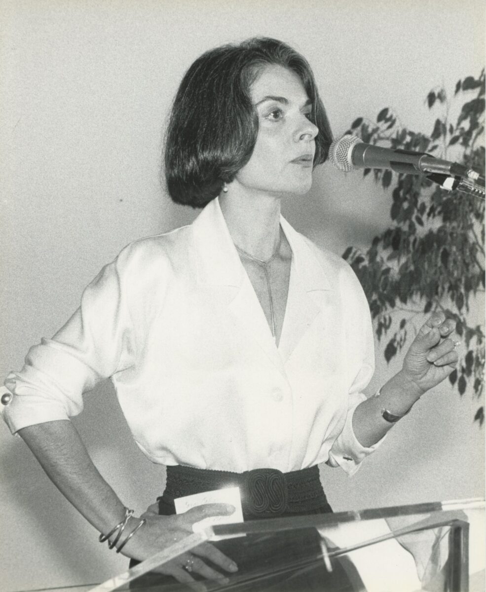 A black and white photo of Cora Cahan speaking into a microphone.