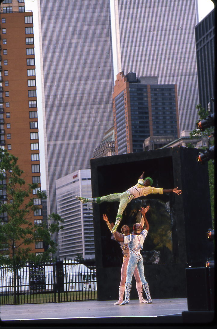 Three male dancers on an outdoor stage dance in multi-patterned costumes. One of the dancers has been thrown in the air and splays his body in an X-shape, while the other two prepare to catch him.