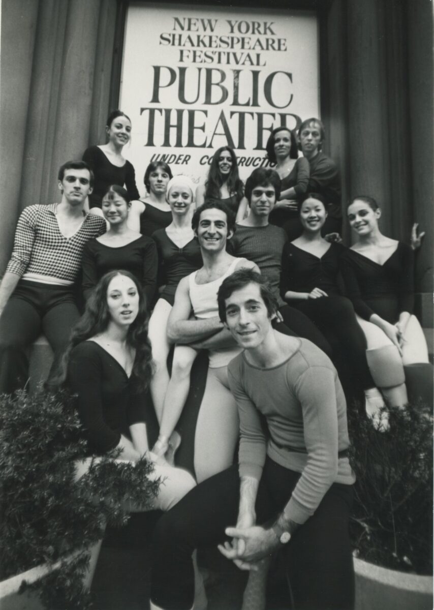 A black and white photo of Eliot Feld and his company dancers, posing together in front of a sign that reads &quot;New York Shakespeare Festival Public Theater.&quot;